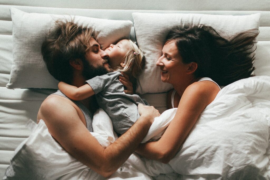 A couple laying down in bed with white sheets embracing a small child.